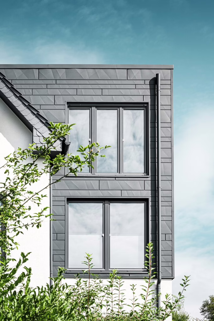 Modernes Wohnhaus mit dunkelgrauer Metallfassade und großen, symmetrisch angeordneten Fenstern auf zwei Etagen. Die Fassade ist mit strukturierten, geometrischen Paneelen verkleidet. Im Vordergrund wachsen grüne Sträucher, darüber blauer Himmel mit vereinzelten Wolken.